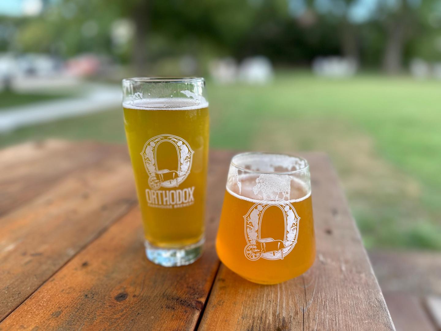 Two glasses of beer on a wooden table outdoors, with a grassy park background.
