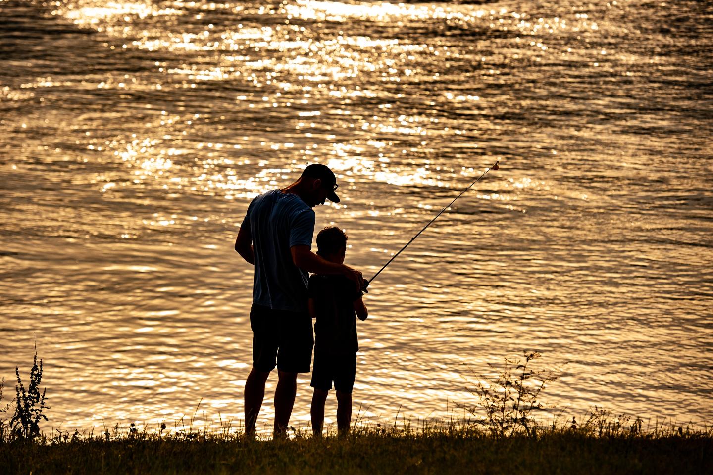 Silhouetted man and child fishing at sunset by a shimmering lake.