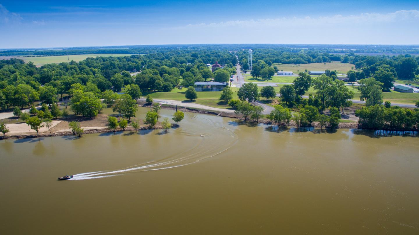 River with a small boat, surrounded by trees and a distant town on a sunny day.