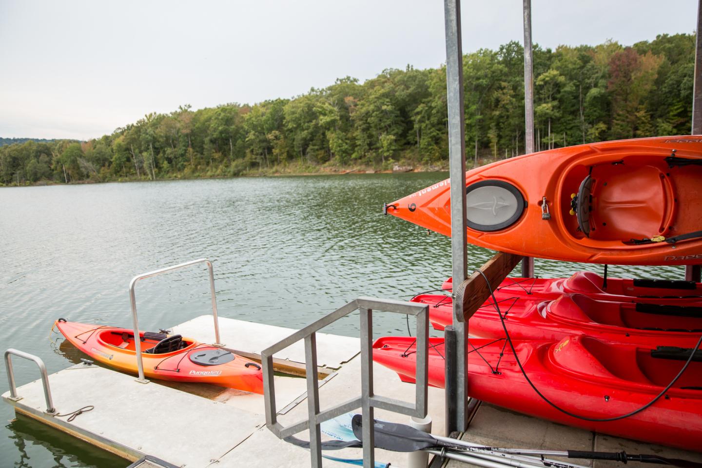 Kayaks stacked on a dock by a calm, tree-lined lake.