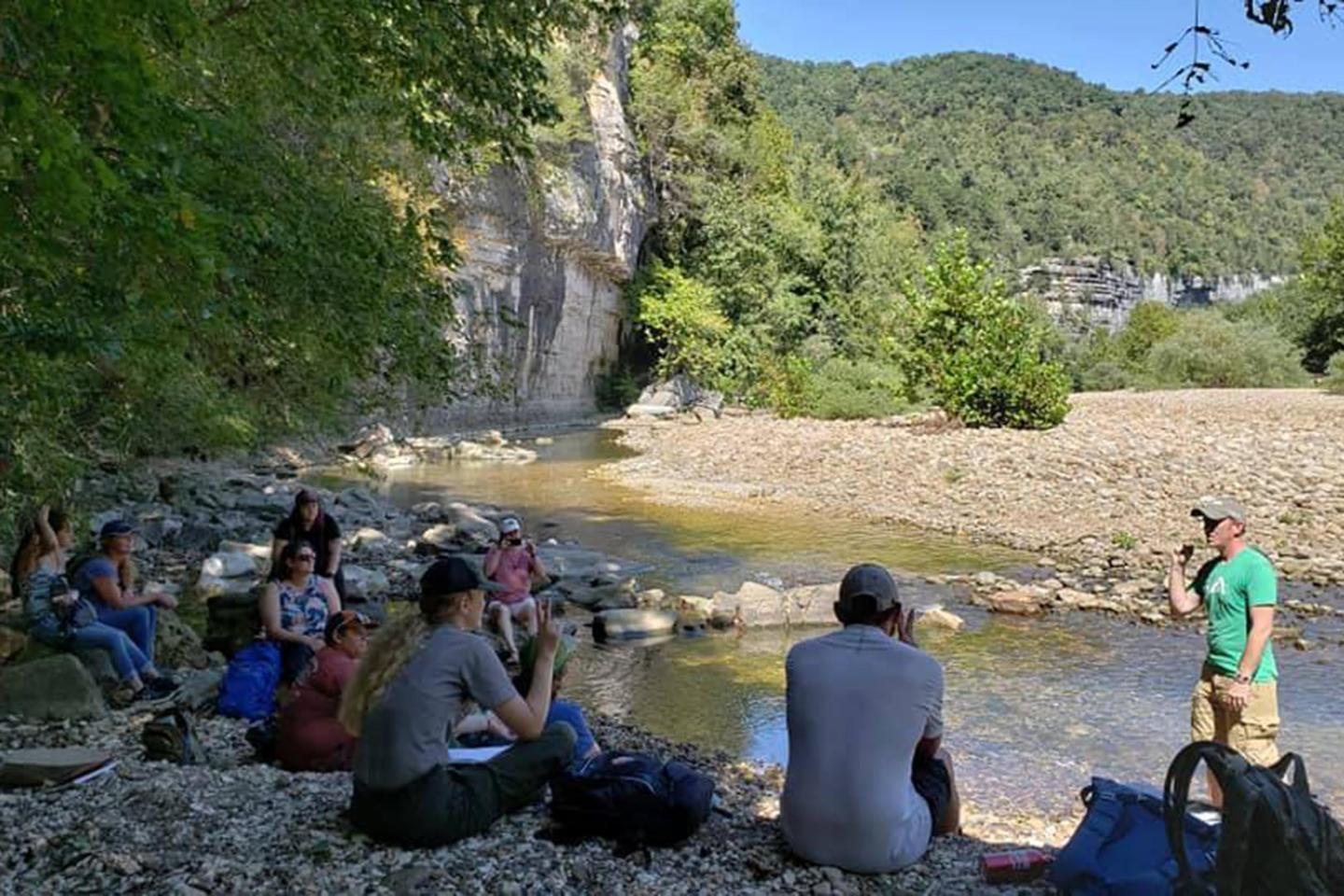 Group sitting by a riverbank, listening to a speaker in a natural, wooded setting.