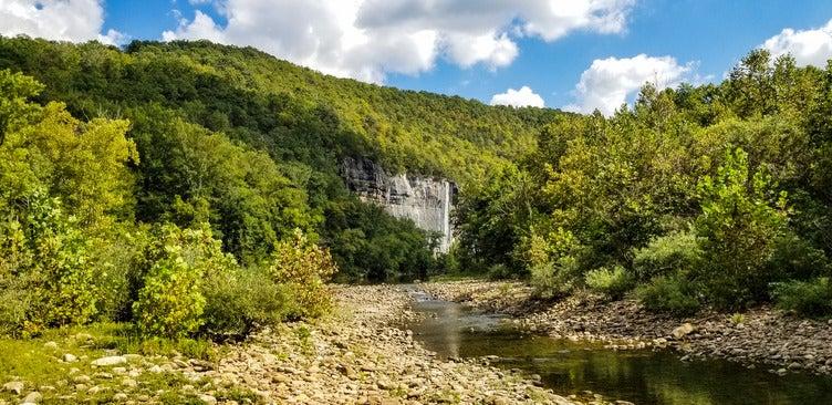 Rocky creek with forested hillside and distant waterfall under blue sky.