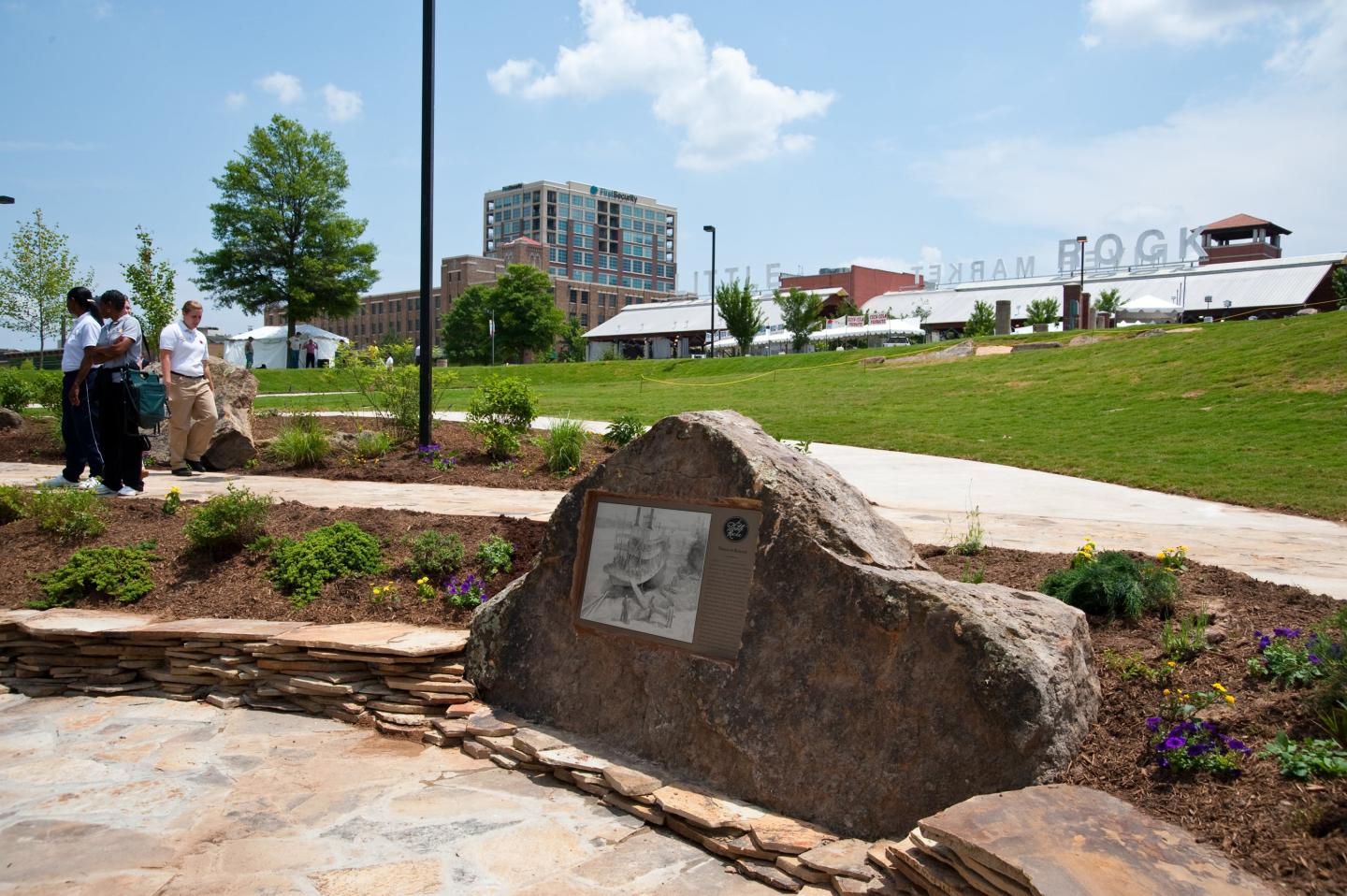 Park path with large rock plaques, people walking, and buildings in the background.