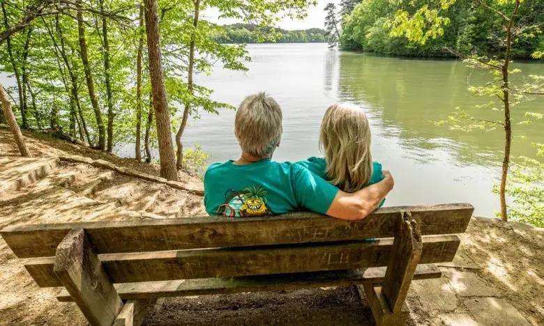 Couple sitting on a bench by a lake, surrounded by trees.