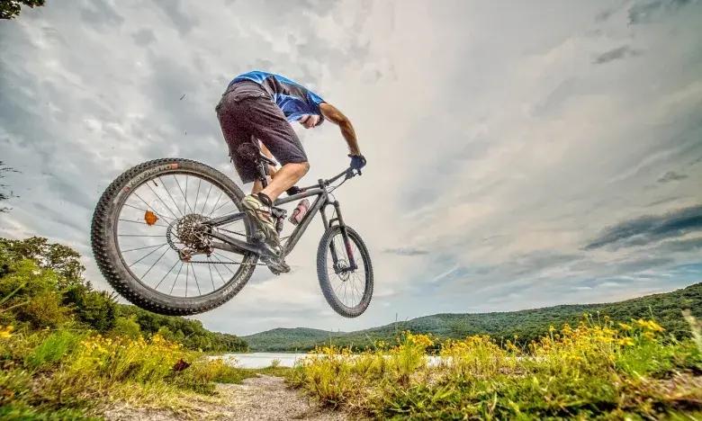 Mountain biker jumps on trail near lake under cloudy sky.