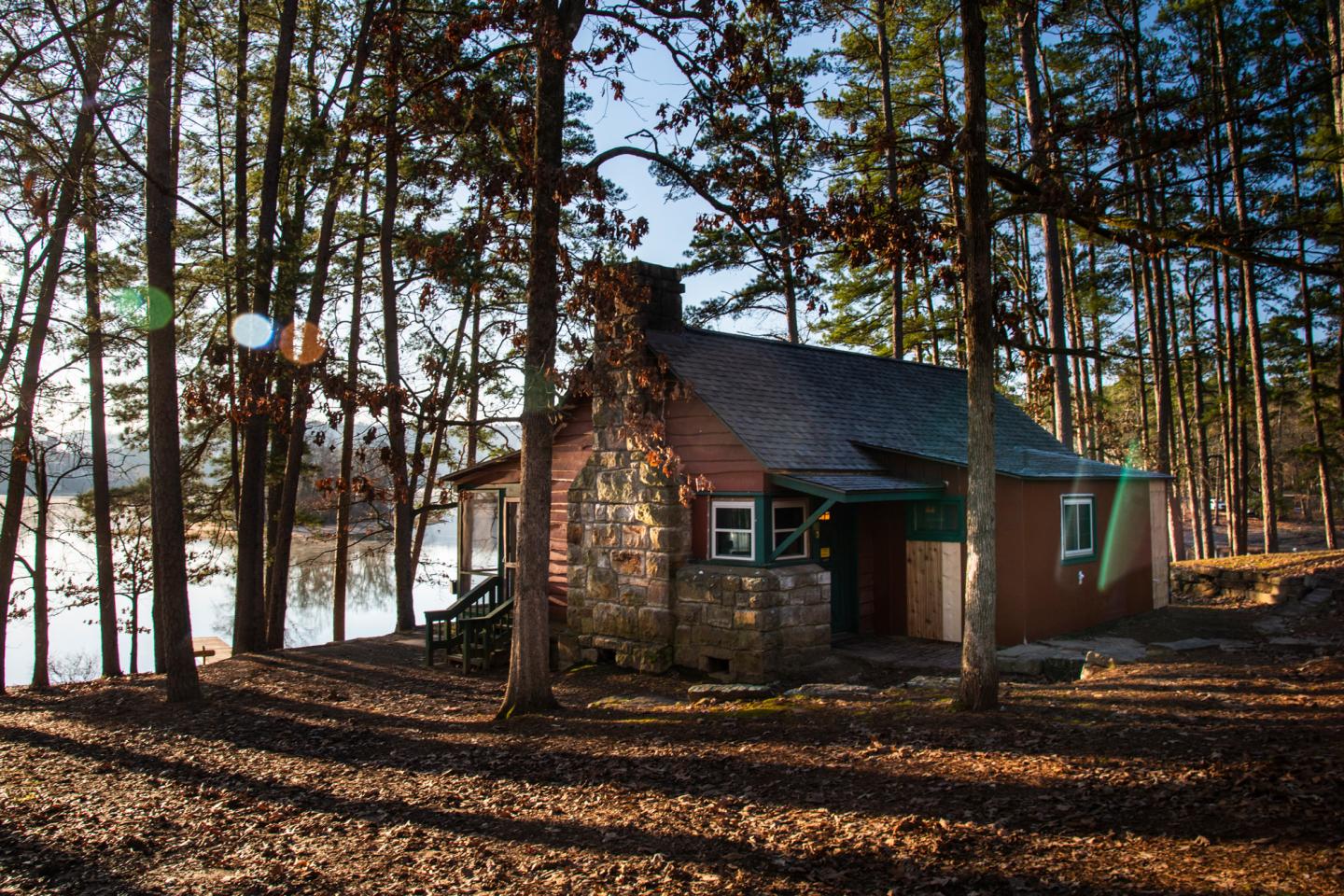 Cozy cabin by a lake surrounded by tall trees in warm sunlight.