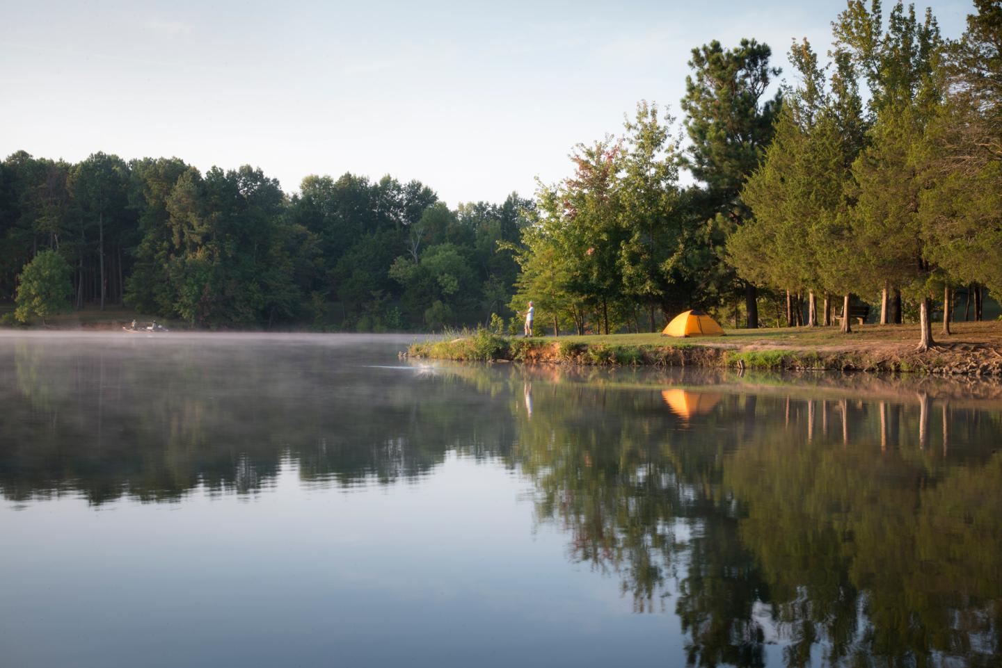 Calm lake with mist, trees, and a tent on the shore at sunrise.
