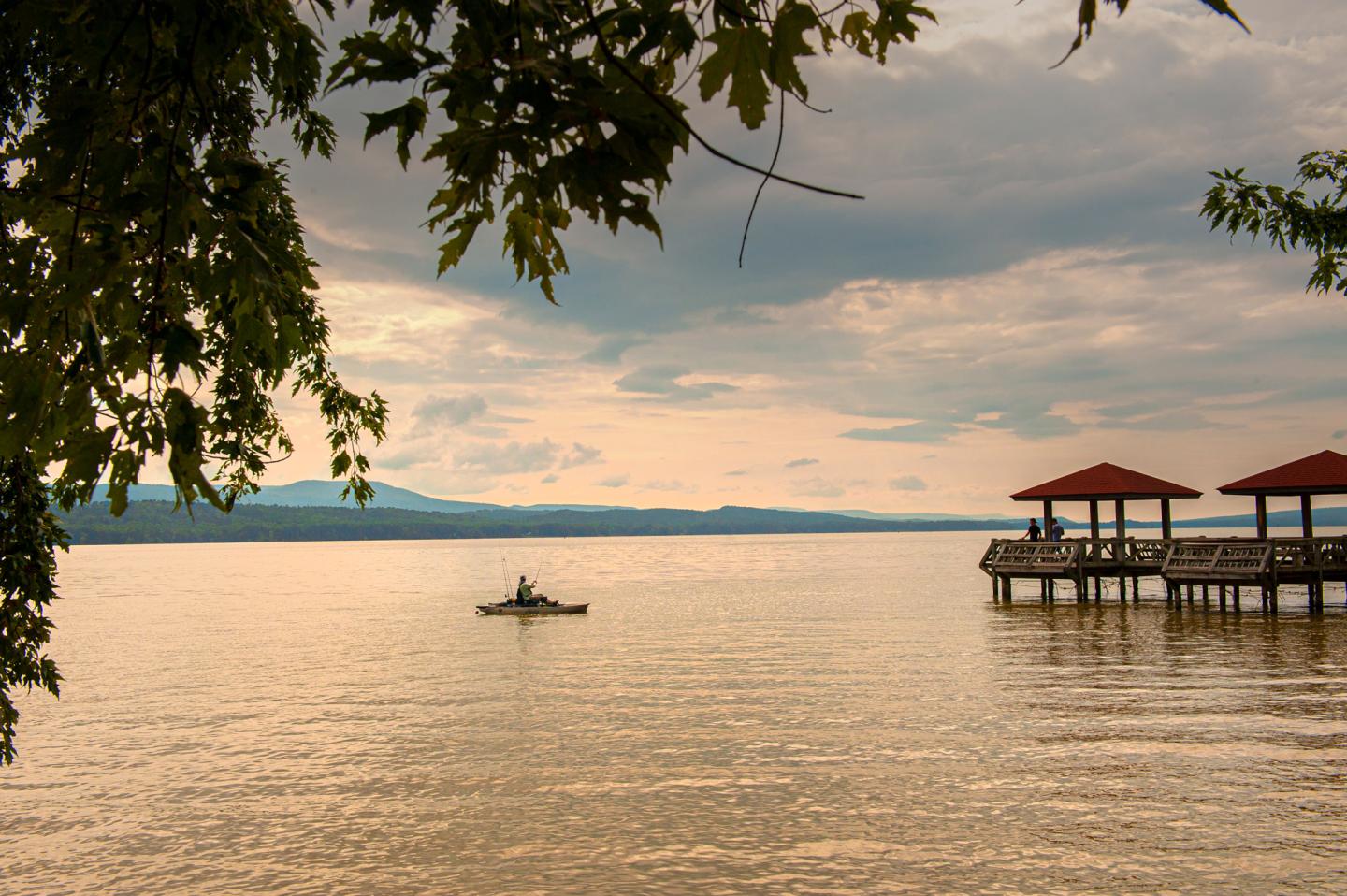 Kayaker on calm lake at sunset near red-roofed gazebos, framed by tree branches.