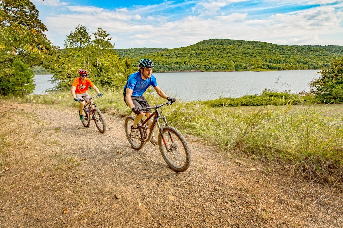 Two cyclists ride along a lakeside trail on a sunny day.