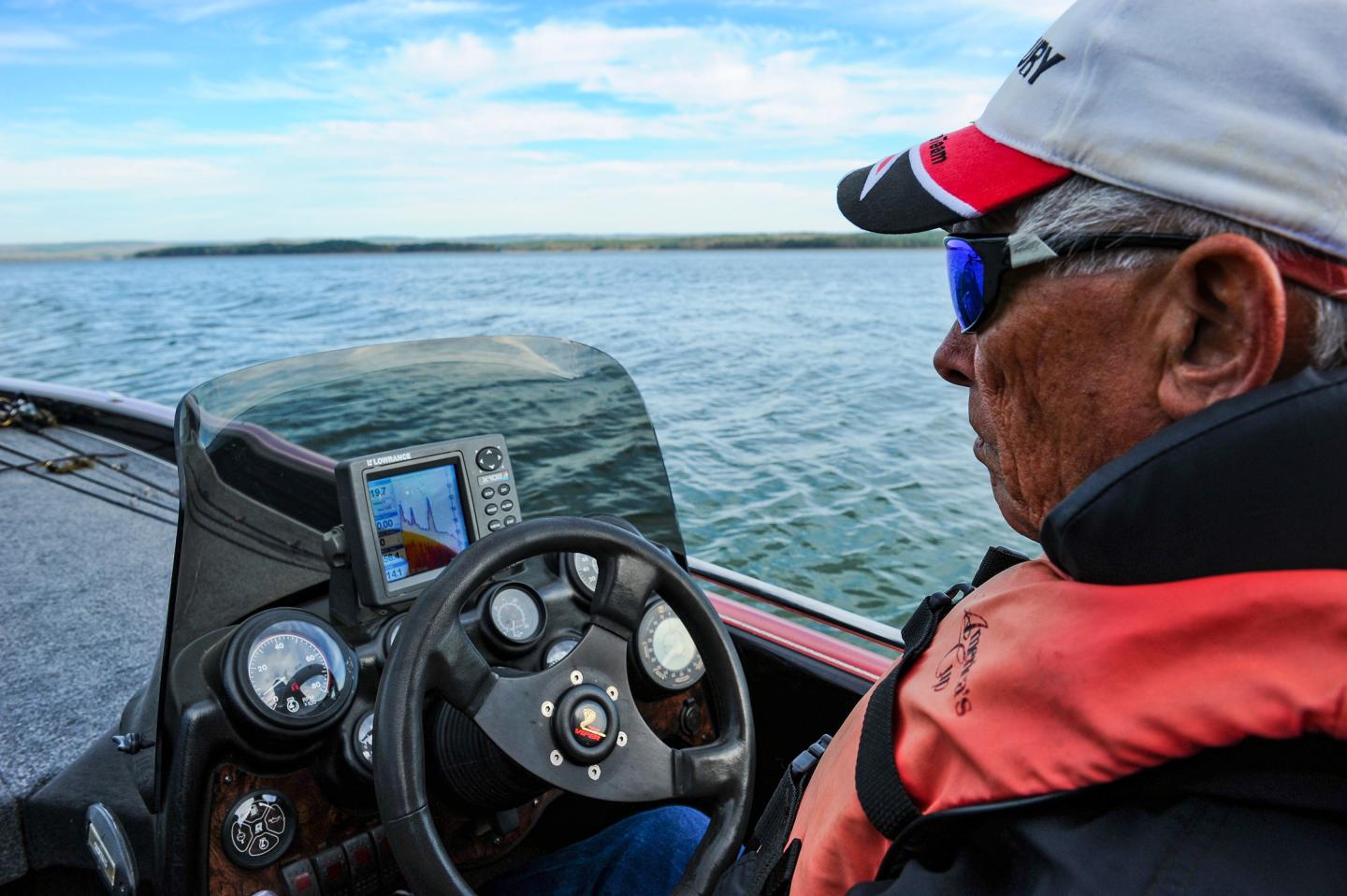 Man steers a boat on a lake, wearing a cap and sunglasses.