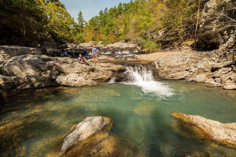 Rocky stream with small waterfall, surrounded by trees and greenery.