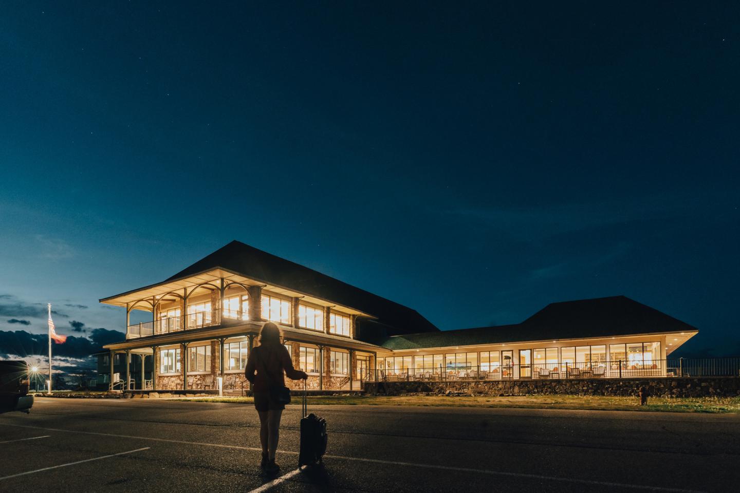 A person with luggage approaches a warmly lit building at night.