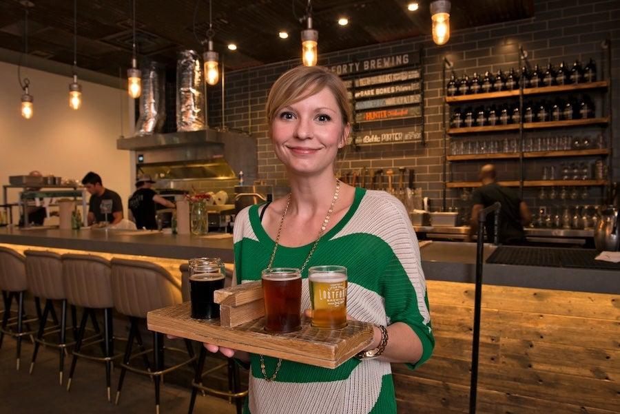 Woman smiling, holding a tray of craft beer samples in a cozy bar.