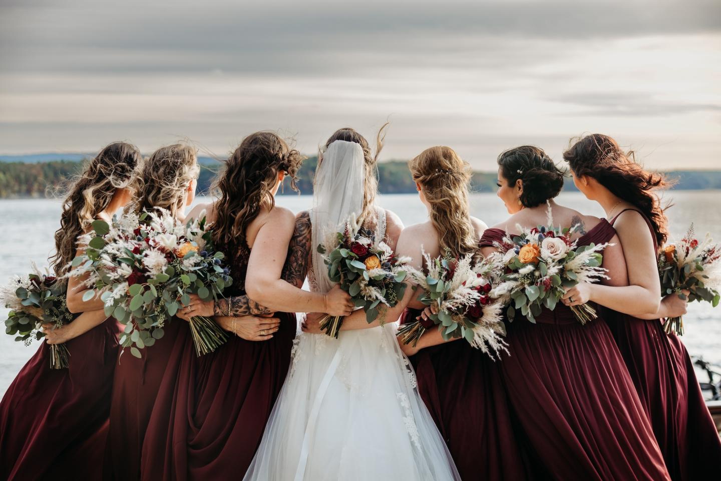Bridesmaids in burgundy dresses and bride facing the water, holding bouquets.