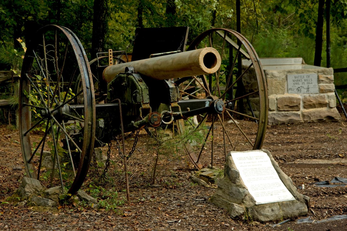 Old cannon on wheels, surrounded by trees with informational plaque.