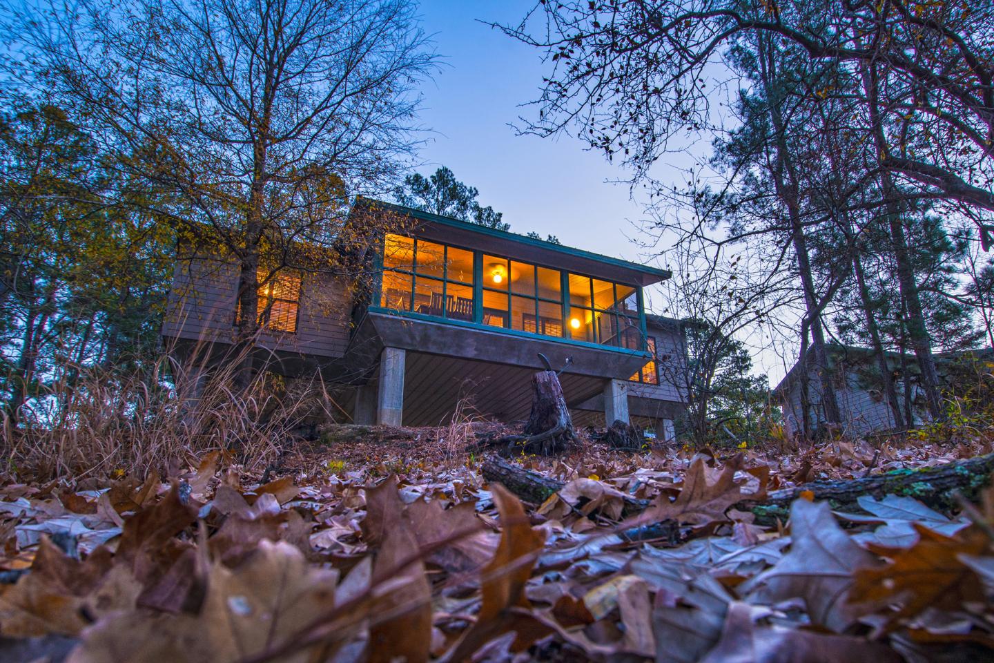 House with large windows, lit warmly at dusk, surrounded by trees and autumn leaves.
