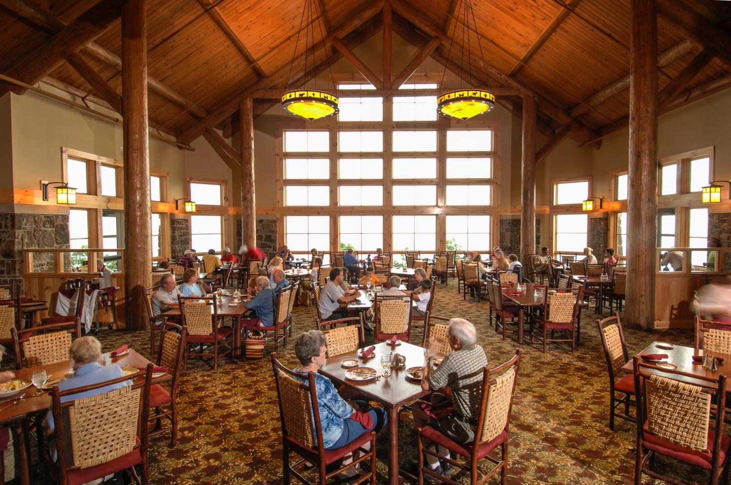 Spacious dining hall with large windows and wooden ceiling, people seated at tables.