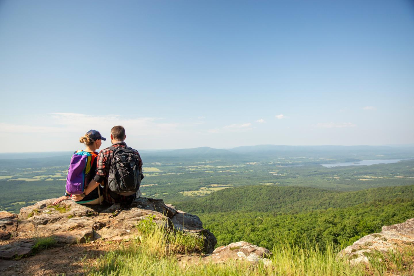 Two hikers with backpacks sit on a cliffside overlooking a vast, green landscape.