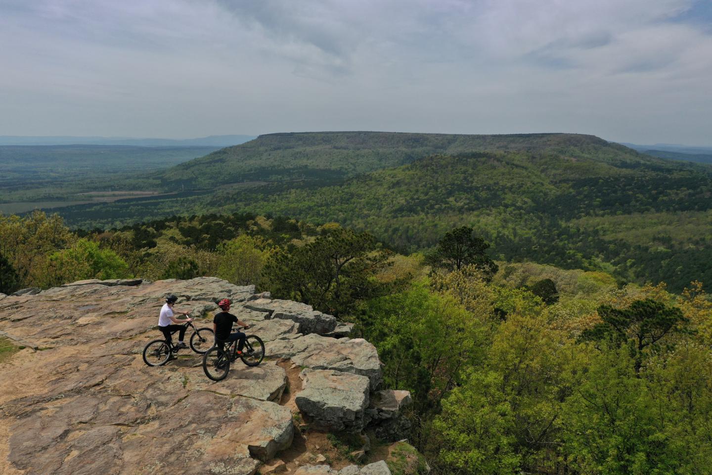 Two cyclists on a rocky overlook, viewing a green forested landscape.