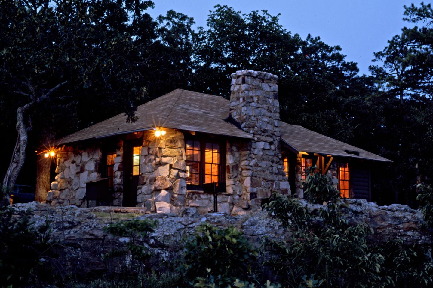 Stone cabin with warm lights, surrounded by trees at dusk.
