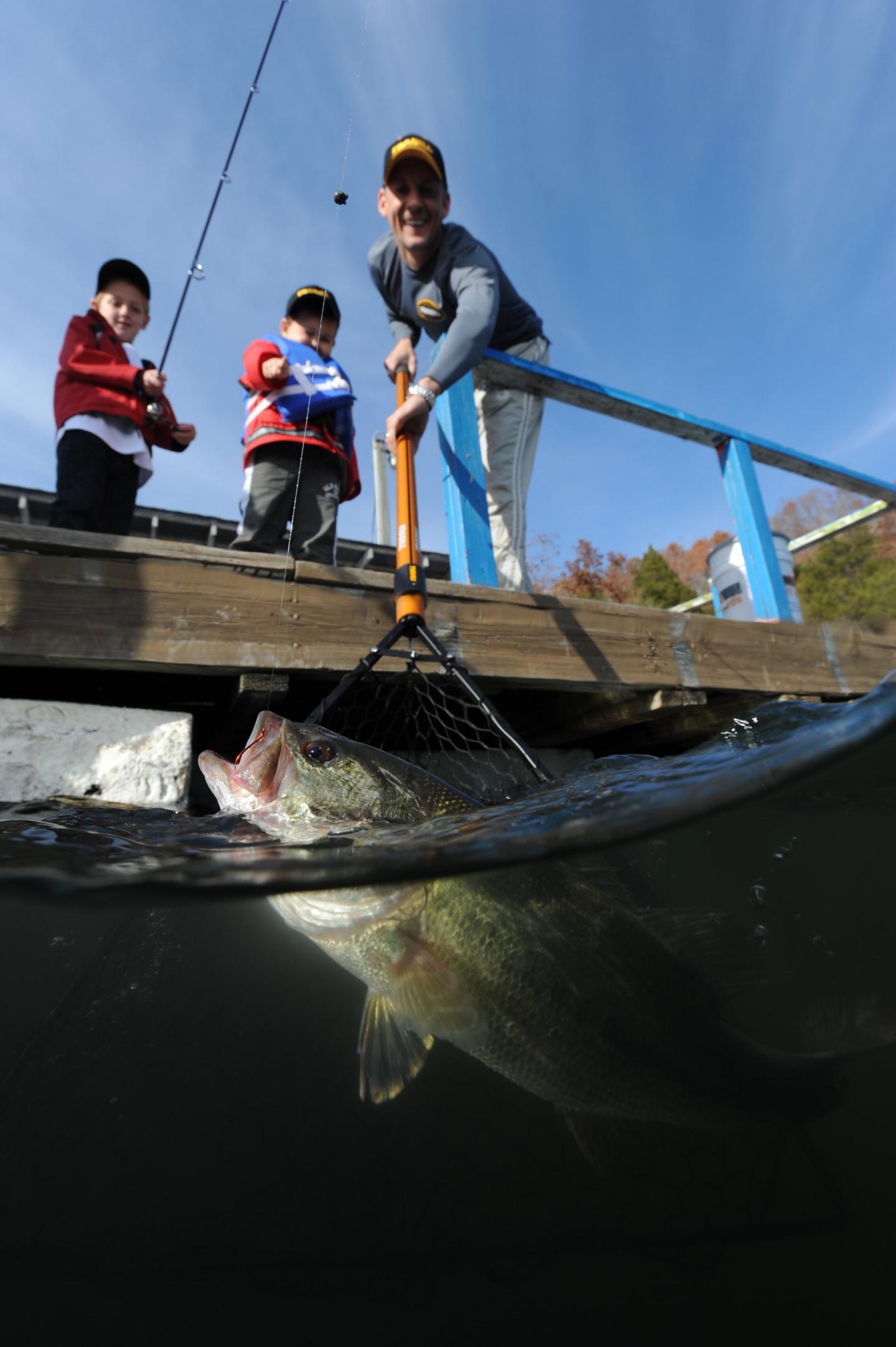 Man catching fish with a net from a dock, two kids watching excitedly.