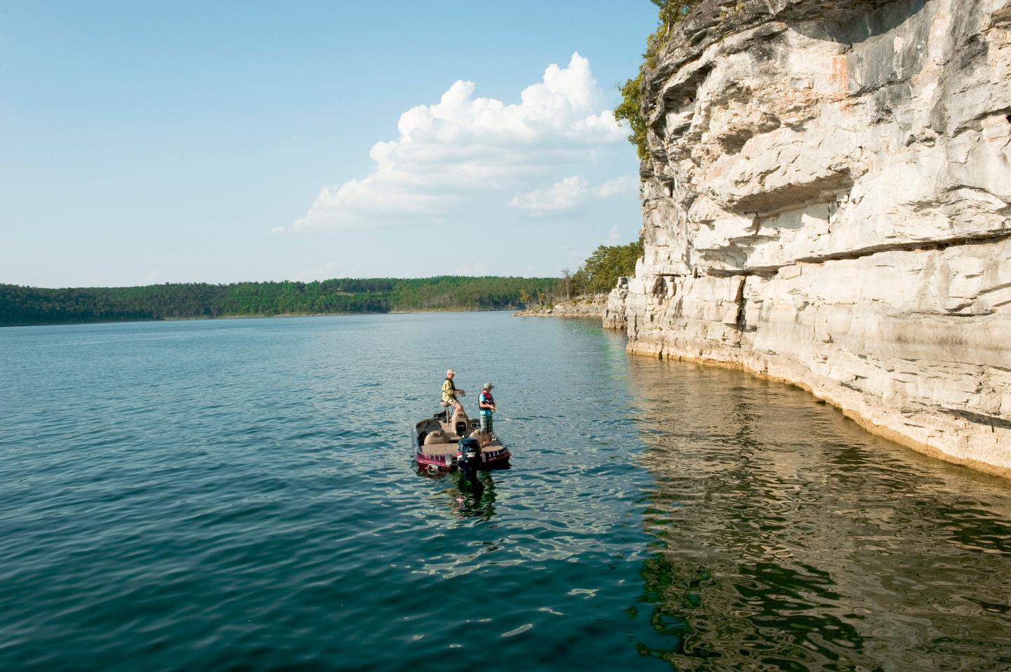 Two people fish from a boat near tall, rocky cliffs under a blue sky.