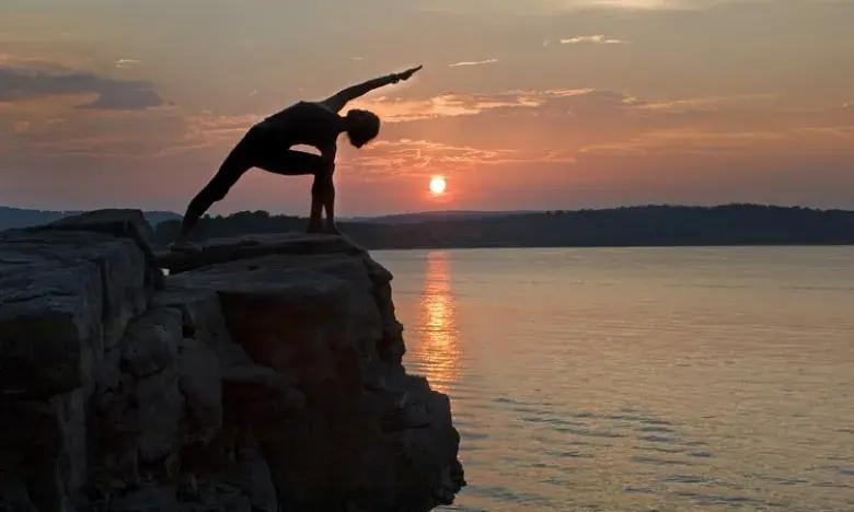 Person doing yoga on a cliff at sunset near a calm lake.