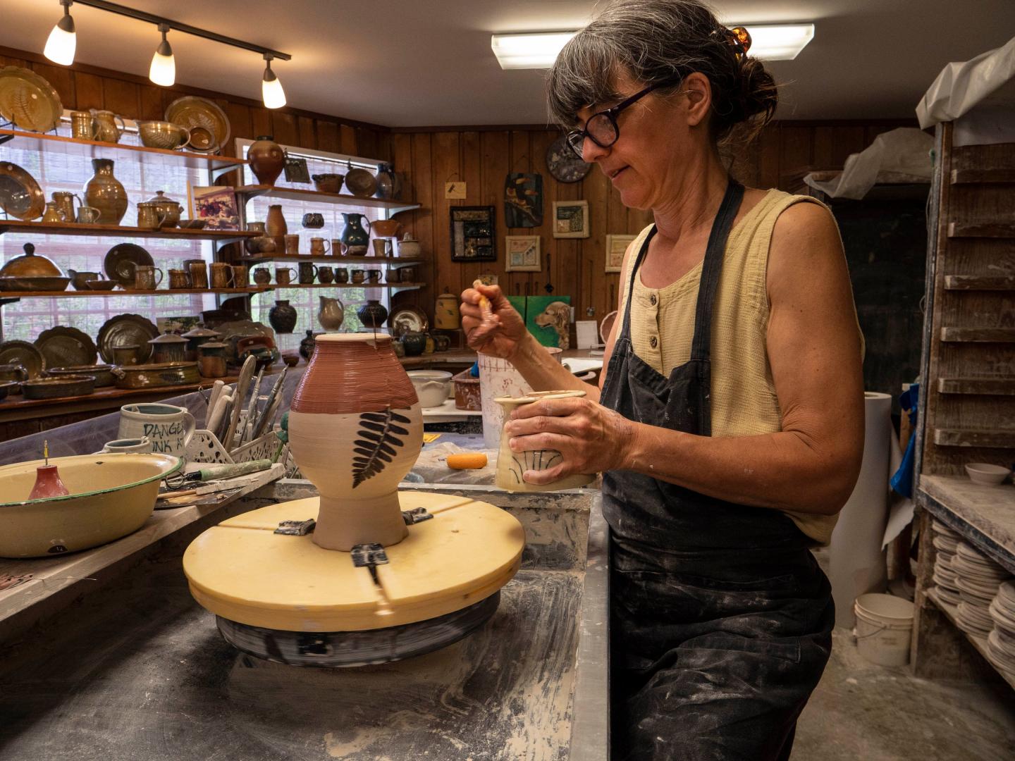 Potter painting a vase in a workshop filled with ceramics.