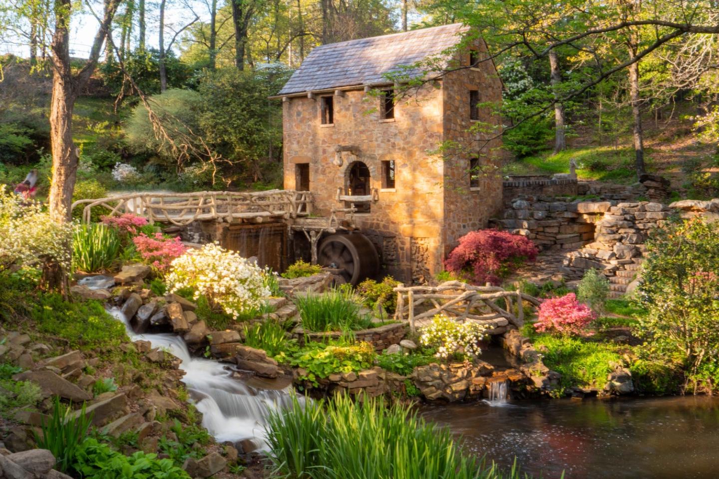 Old stone mill by a stream, surrounded by lush greenery and colorful flowers.