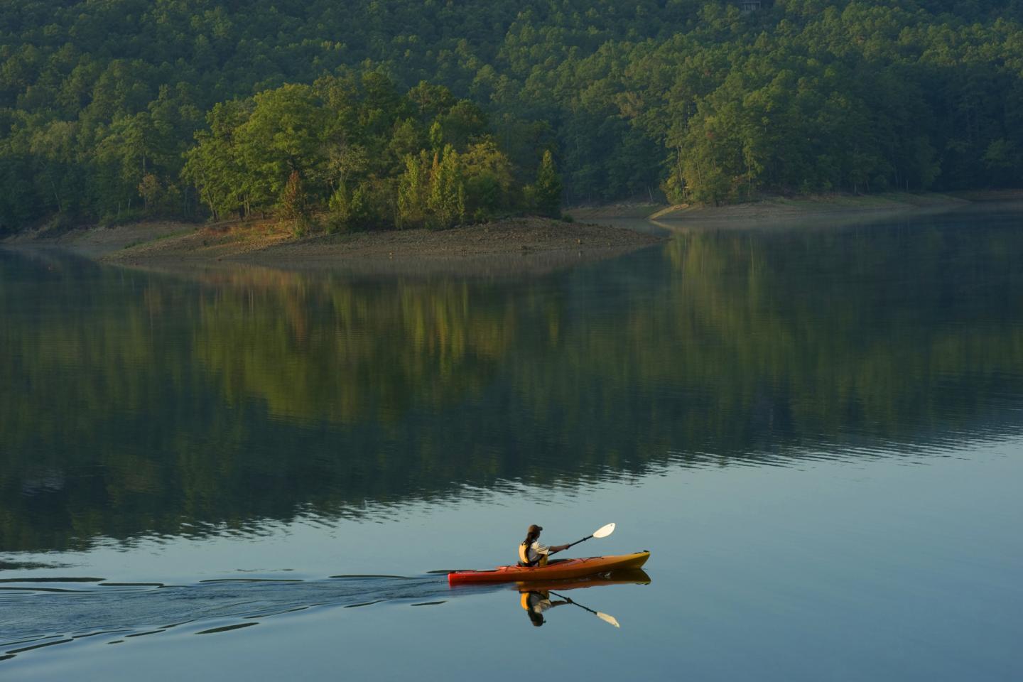 Kayaker on calm lake with forested shoreline reflecting in water.
