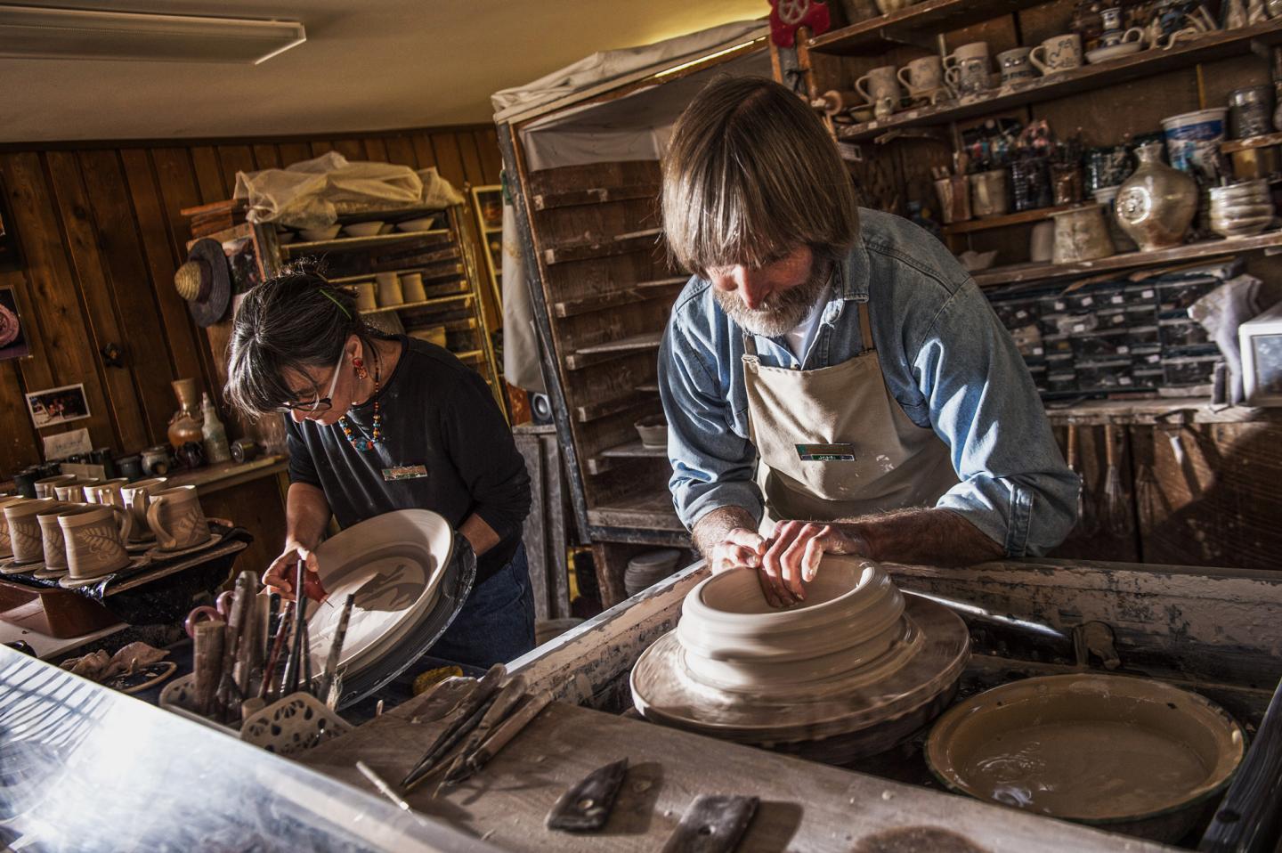 Two people in a pottery workshop, crafting clay pots on wheels.