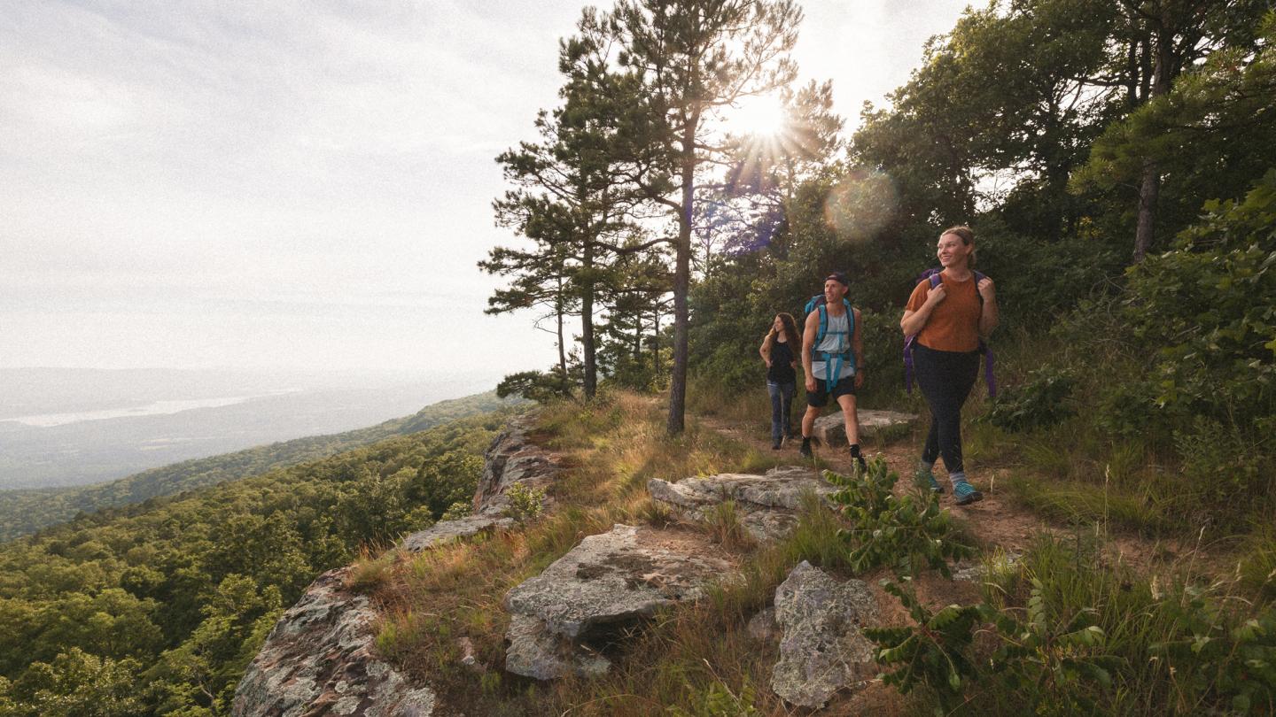 Three hikers on a forested trail at sunrise, with distant hills visible.