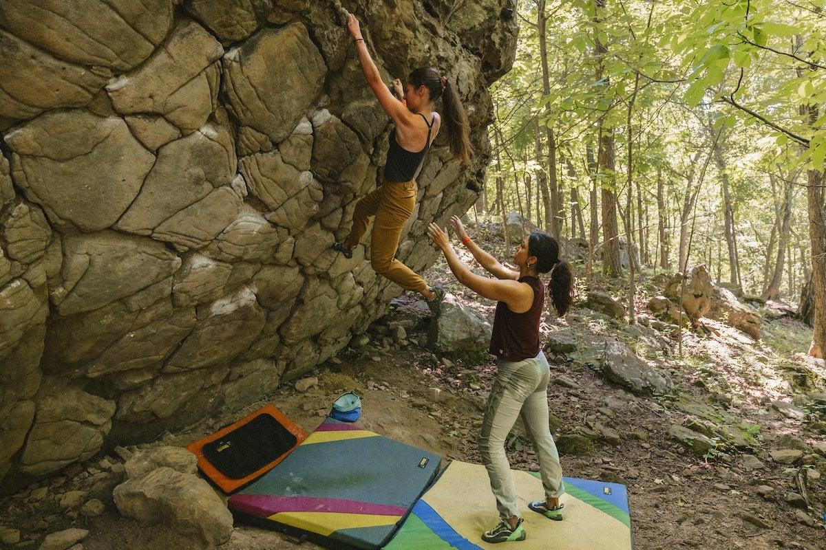 Climber scaling a boulder in a forest, with a spotter and crash pads below.