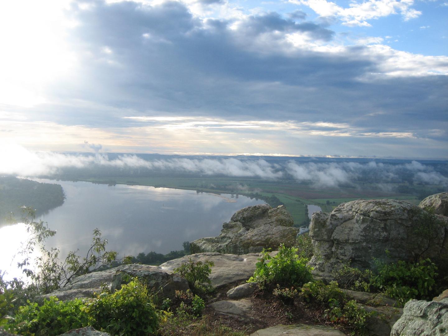 Sunrise over a river with rocks and lush landscape.