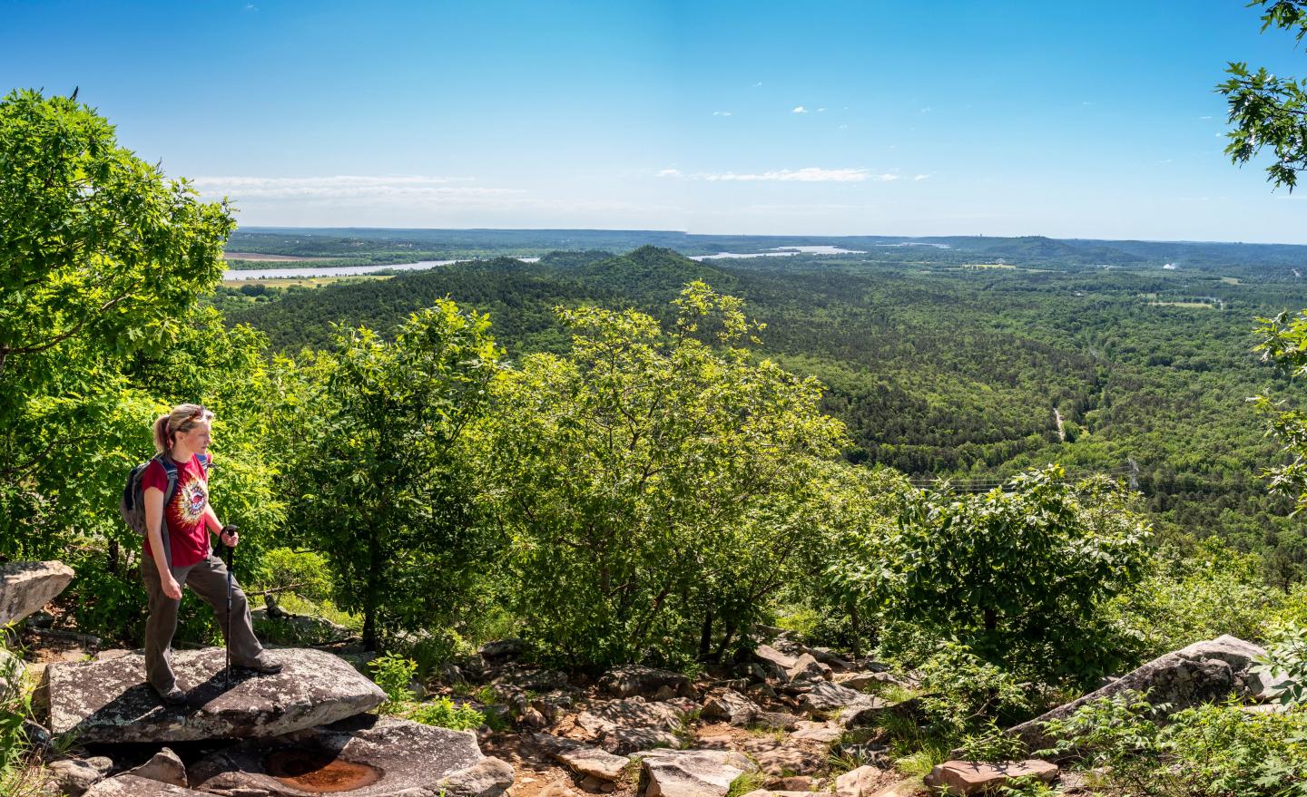 Hiker admiring a vast green landscape from a rocky overlook under a clear sky.