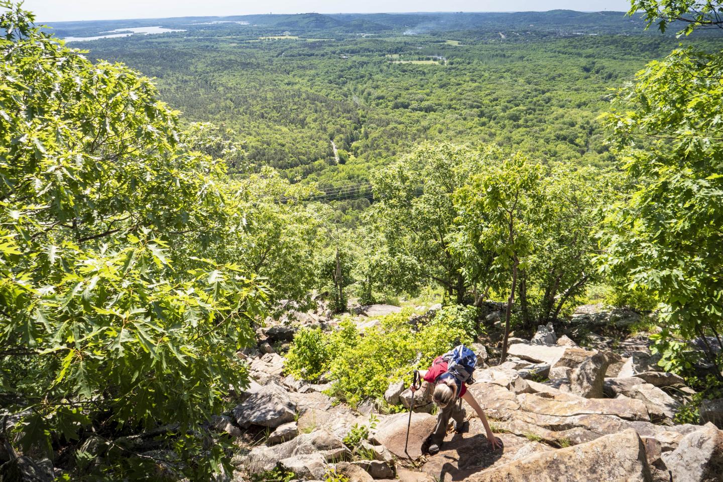 Hiker climbs rocky trail overlooking lush green forest and distant hills.