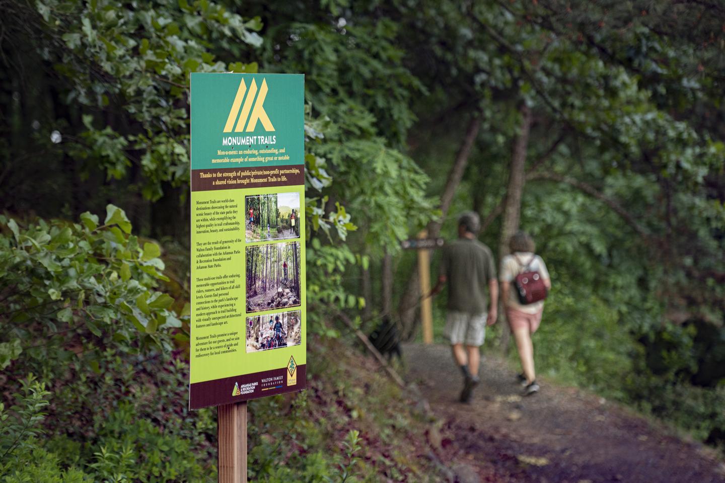 Trail sign beside two hikers on a forest path.