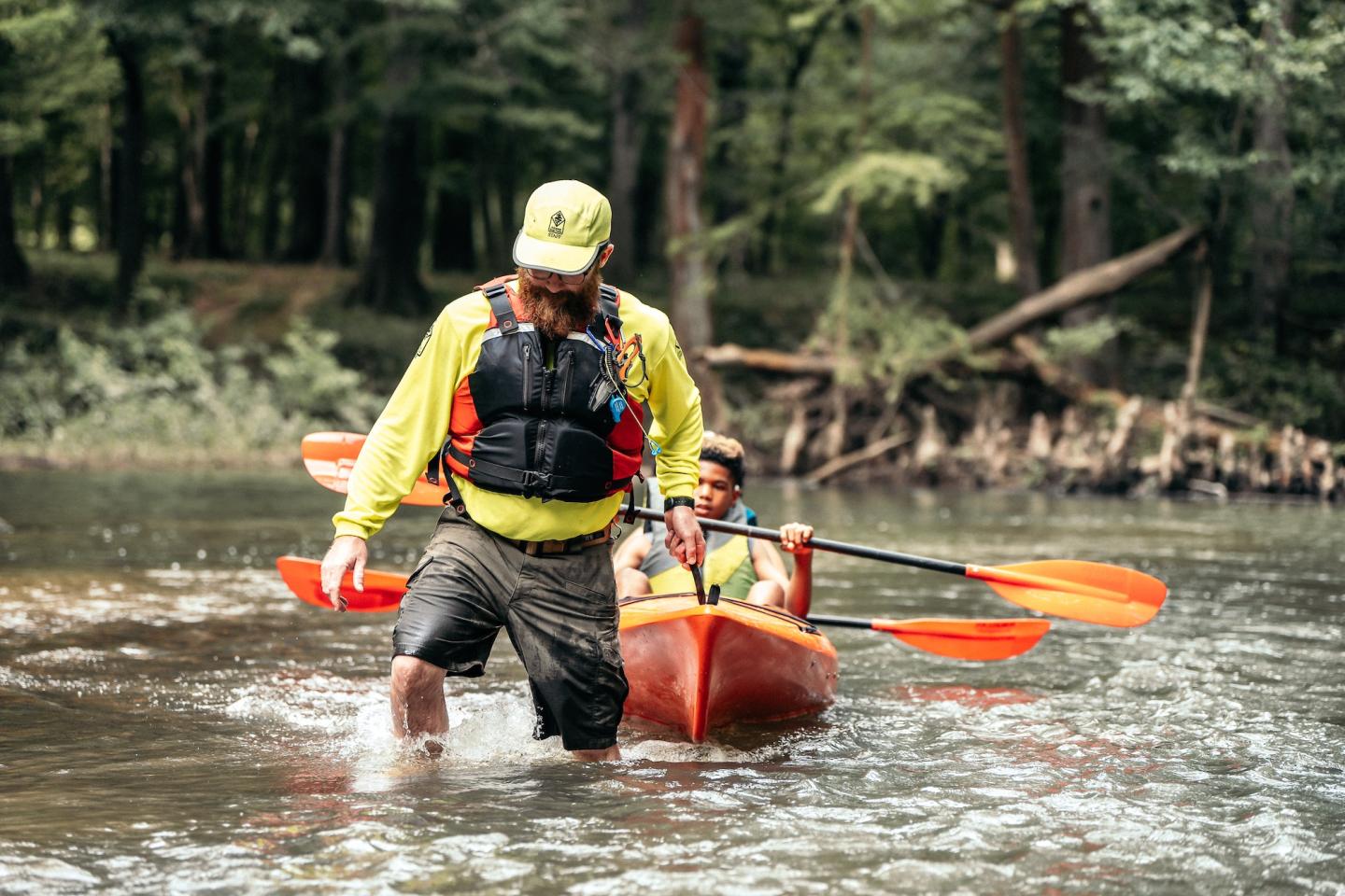 Man walking in a river guiding a red kayak with another person seated inside.
