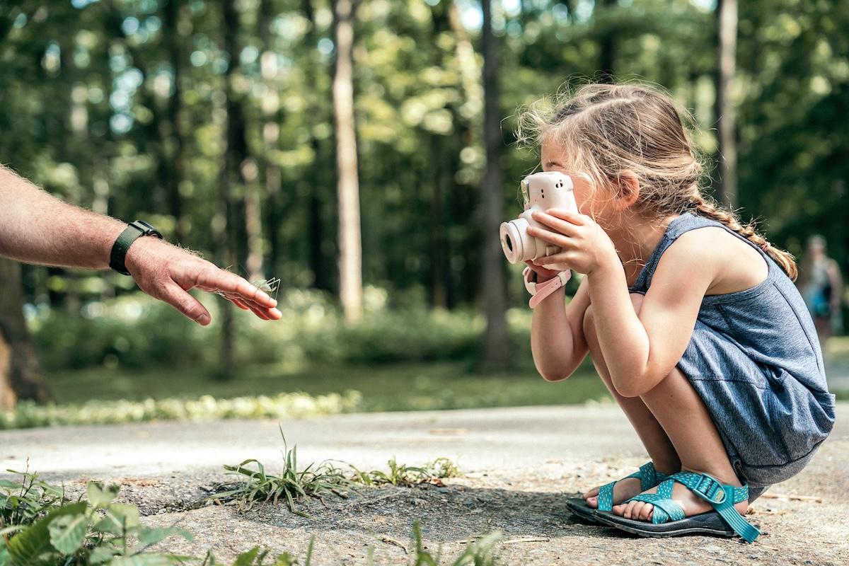 Child crouching with a camera, hand reaching out, sunny forest background.