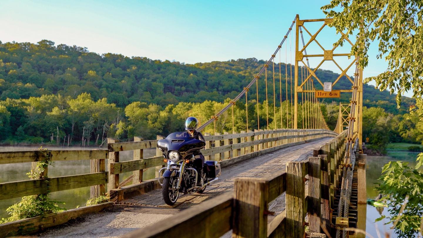 Motorcyclist on a bridge with scenic, tree-covered hills in the background.