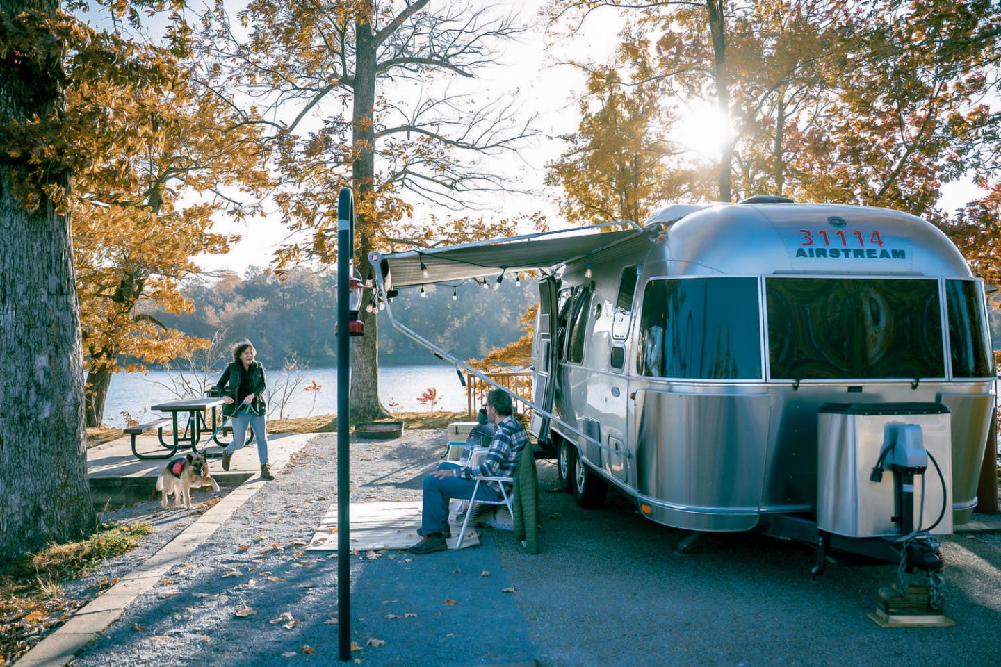 Silver camper parked by a lake in autumn, sun shining through trees.