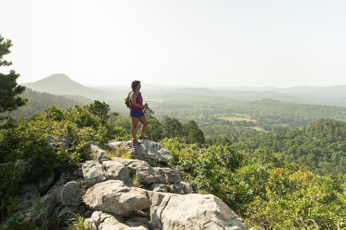 Hiker on rocky cliff overlooking vast green landscape and distant mountains.