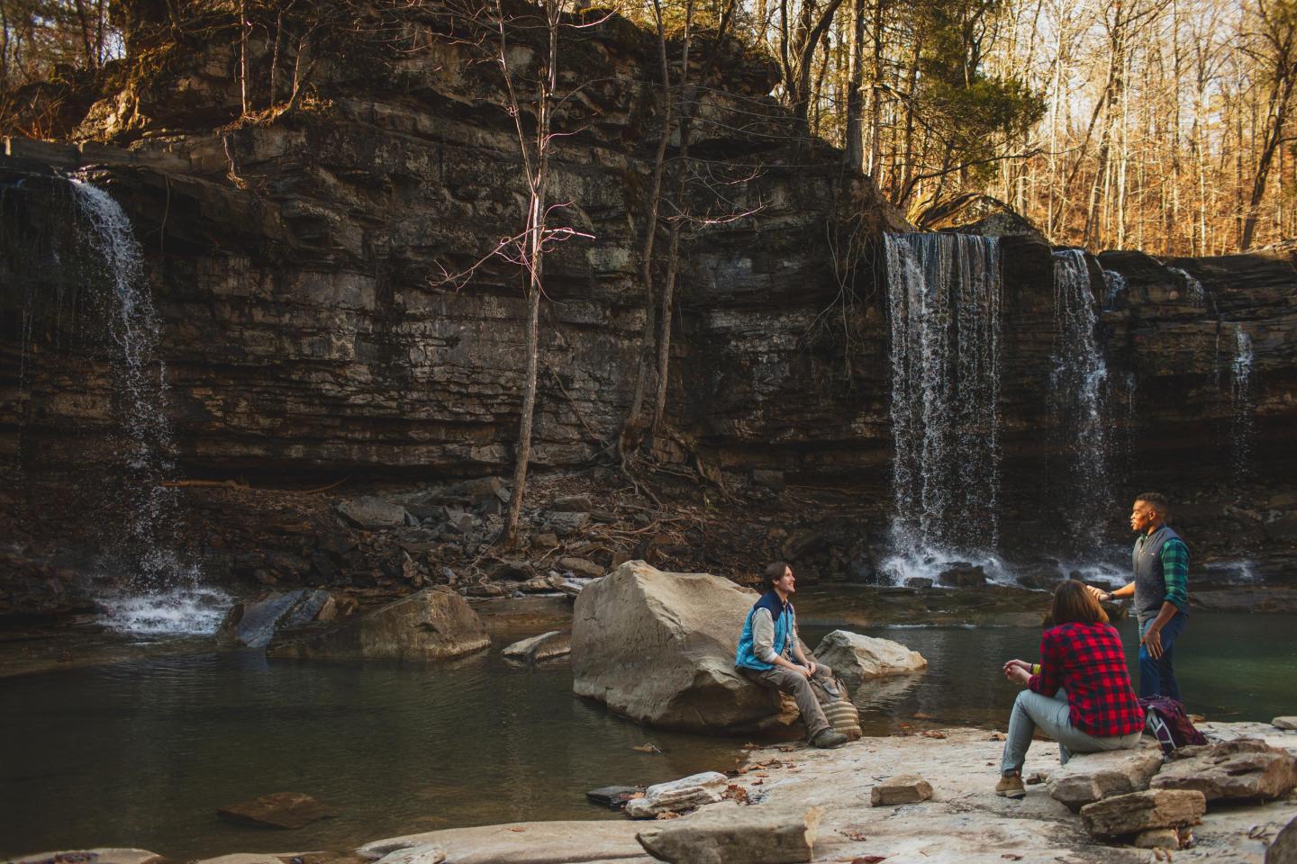 Waterfall scene with four people sitting and standing on rocks, surrounded by trees.
