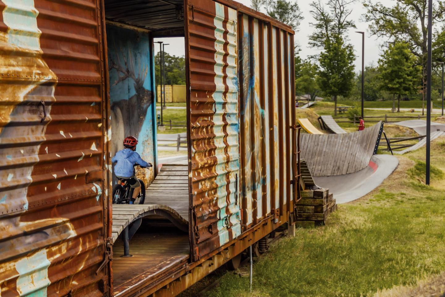 Child cycling through a rusty train car in a park with ramps and trees.