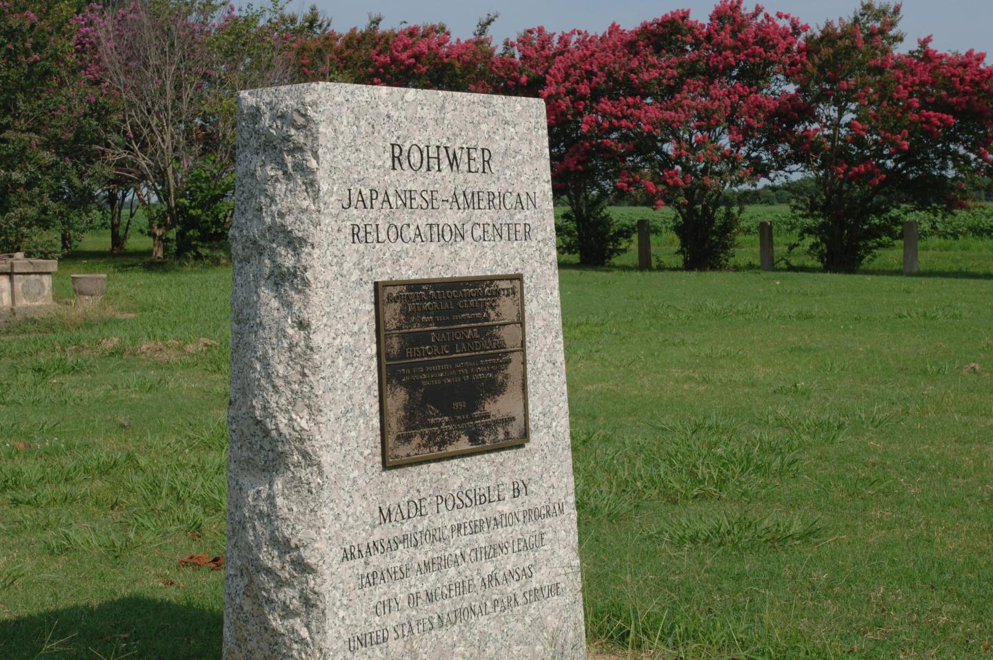 Stone memorial with plaque in a grassy area, pink flowering trees in the background.
