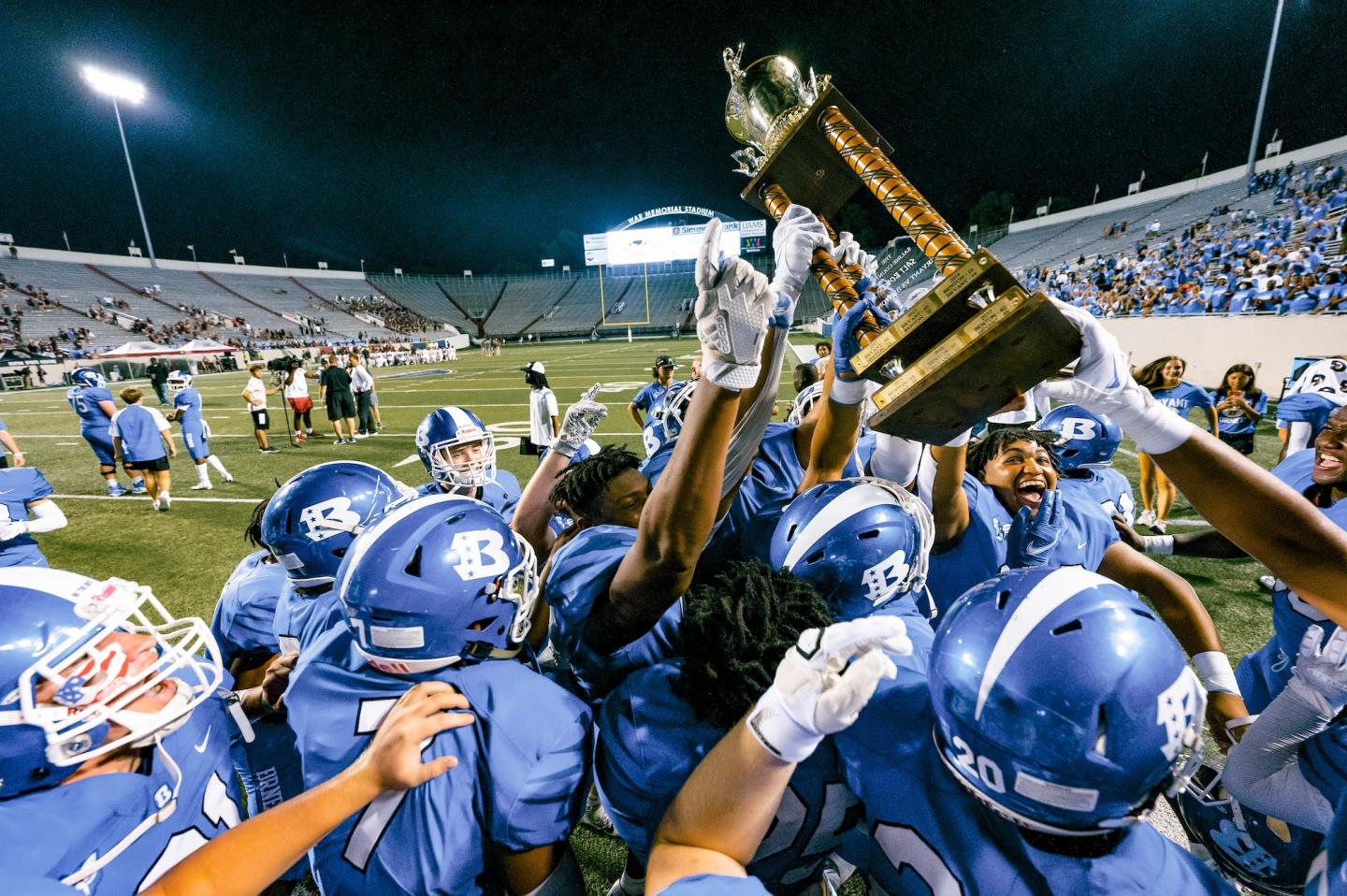Football team in blue jerseys celebrating with a trophy on a field at night.