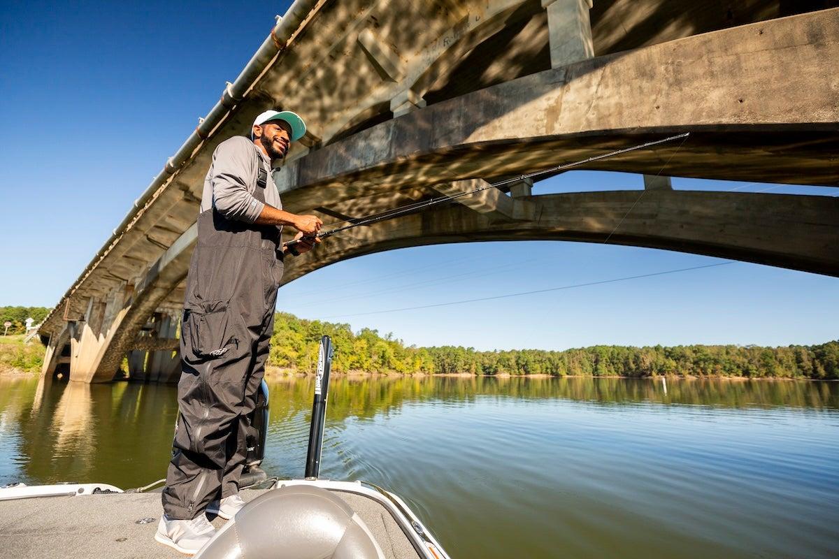 Under a bridge, a person fishes from a boat on a calm lake.
