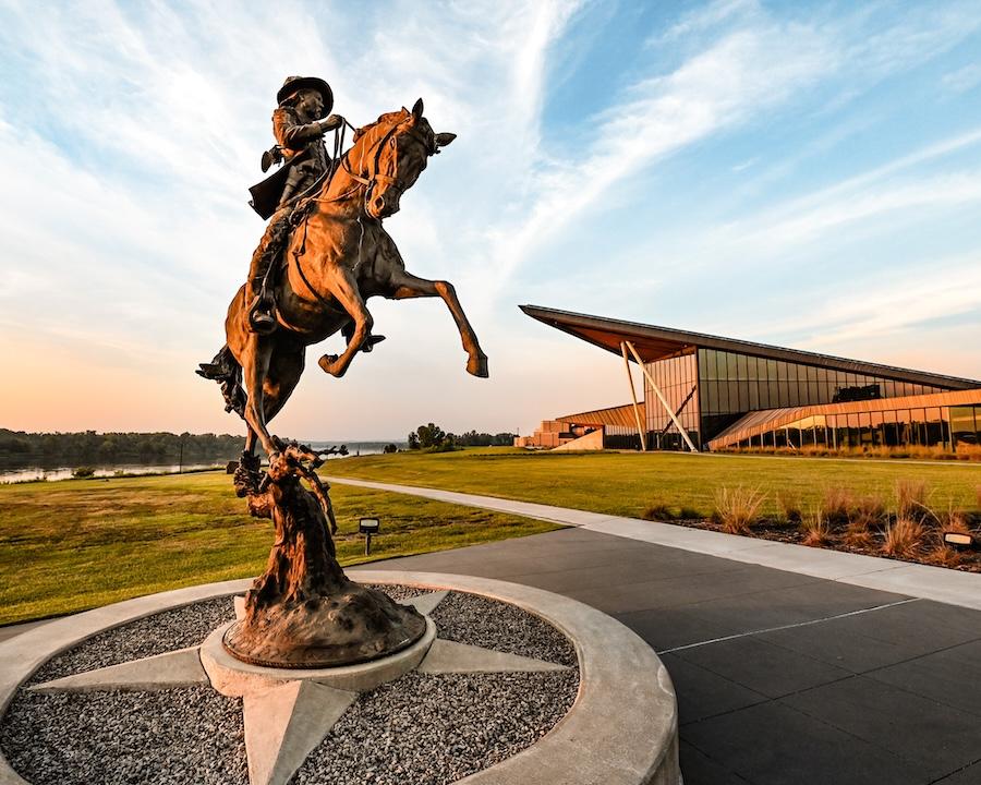 Bronze horseman statue rearing, with modern building and sunset sky background.