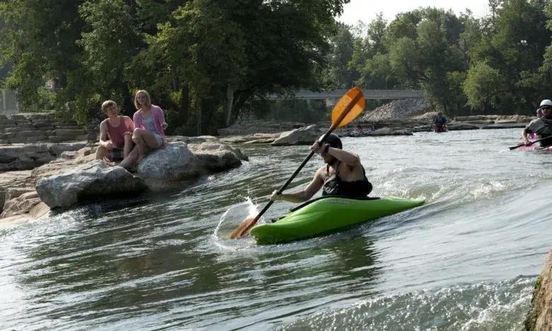 Kayaker paddling swiftly down a river, with people watching from the rocks.