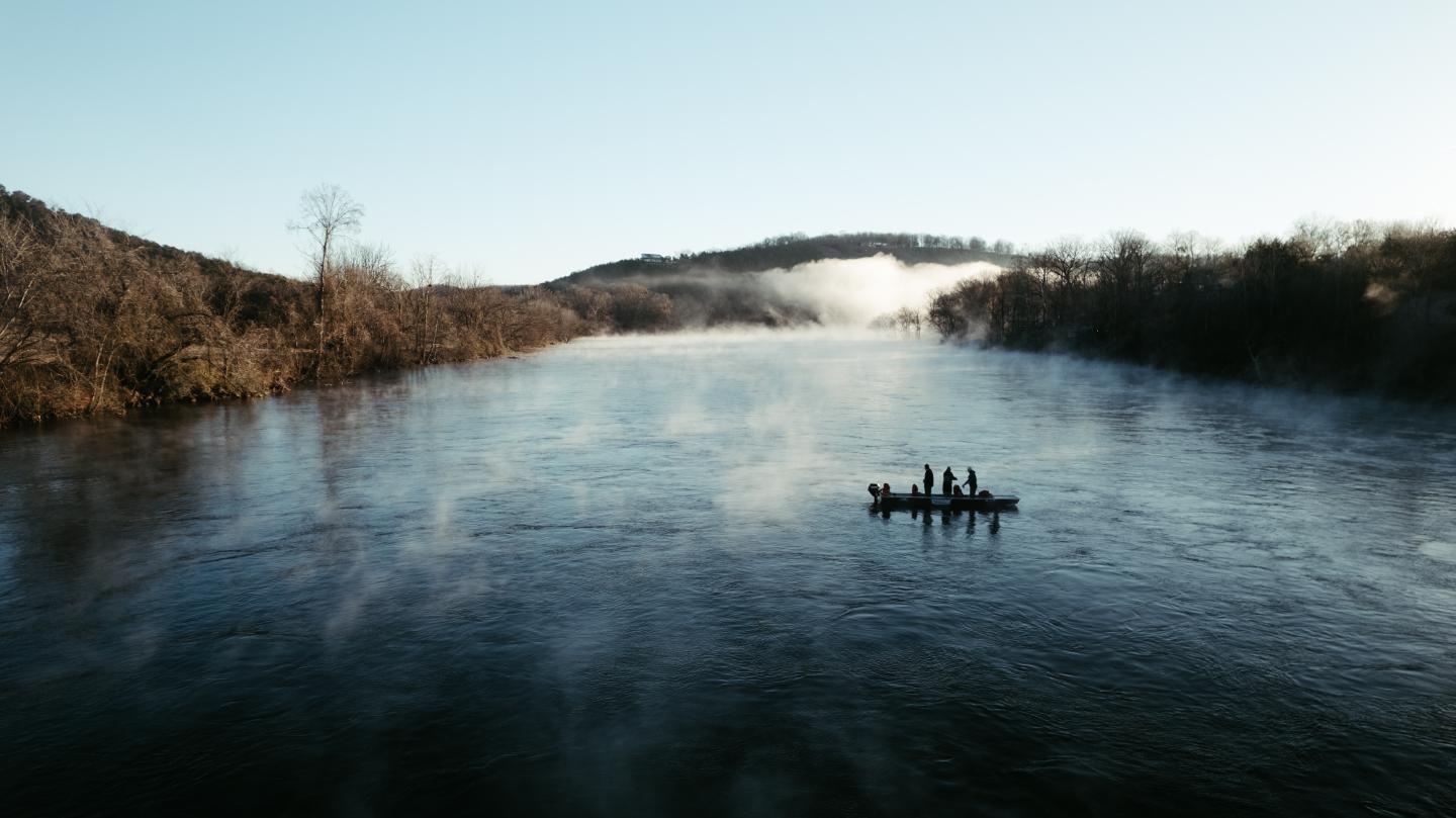 Canoe with people on a misty river, surrounded by hills and trees.